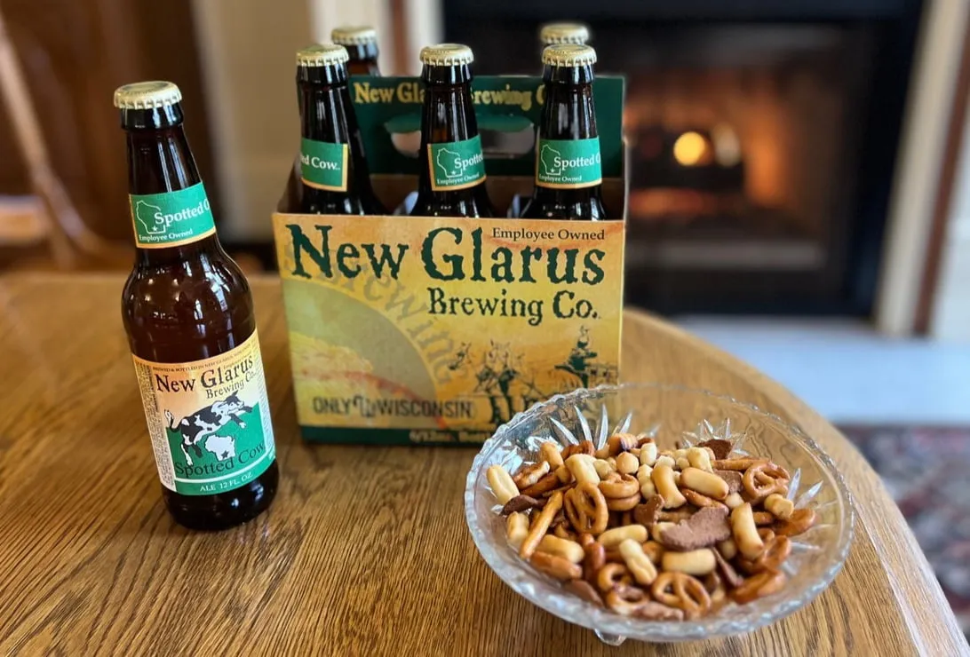 six pack of Wisconsin beer on a wooden table with a bowl of snack mix
