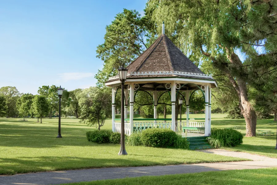 A classic white wooden gazebo with a dark shingled roof sitting in the middle of a spacious, manicured green park surrounded by lush trees and lampposts