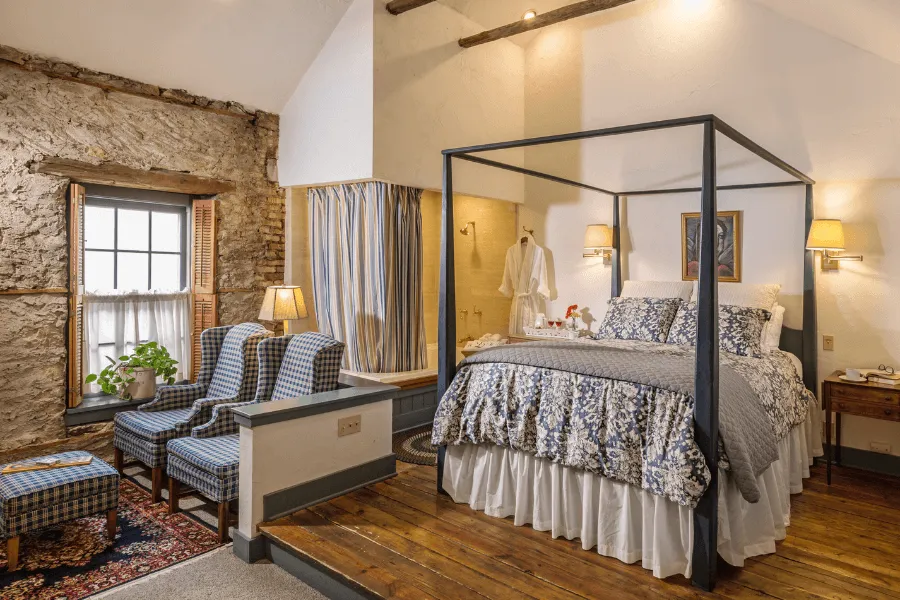 A cozy, rustic hotel guest room featuring a four-poster canopy bed with blue and white floral bedding, an exposed stone wall, and two blue plaid armchairs