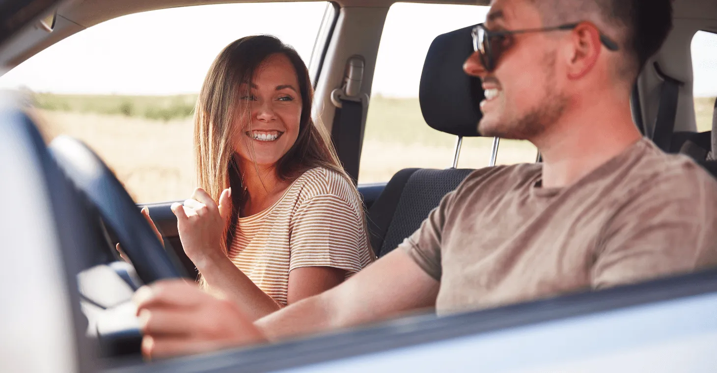 Close-up of a happy couple laughing and talking inside a car during a sunny daytime road trip, with the woman looking warmly at the driver