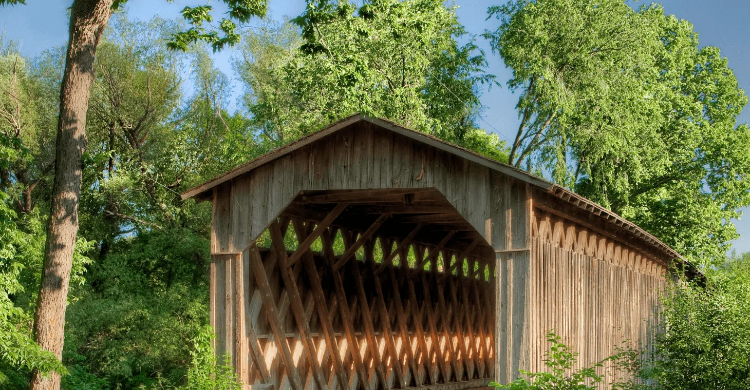A rustic, historic wooden covered bridge surrounded by dense green trees and foliage under a bright blue sky