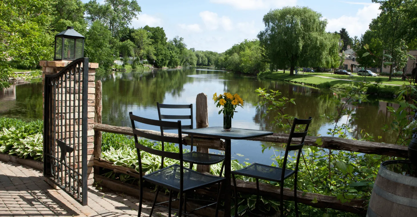 peaceful outdoor patio setting with a black metal table and chairs, a vase of yellow flowers, and a brick gateway overlooking a calm, reflective river lined with trees